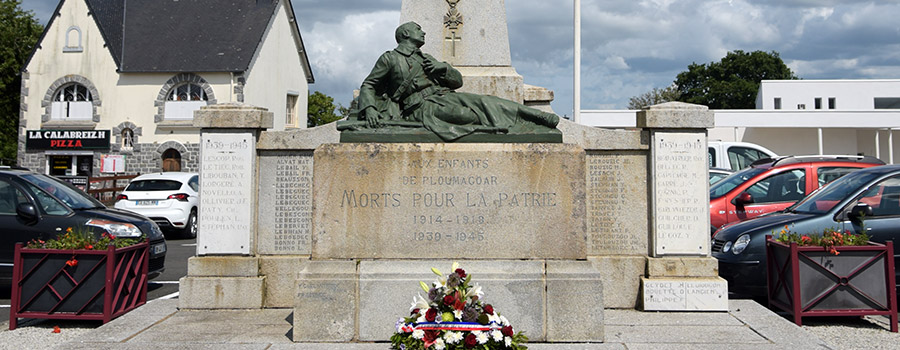 Gros plan sur le monument aux morts de Ploumagoar. Une gerbe de fleurs au premier plan avec un soldat allongé derrière
