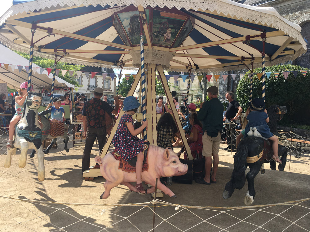 Des enfants montent sur un manège ancien décoré d’animaux, entourés de familles et de fanions colorés.