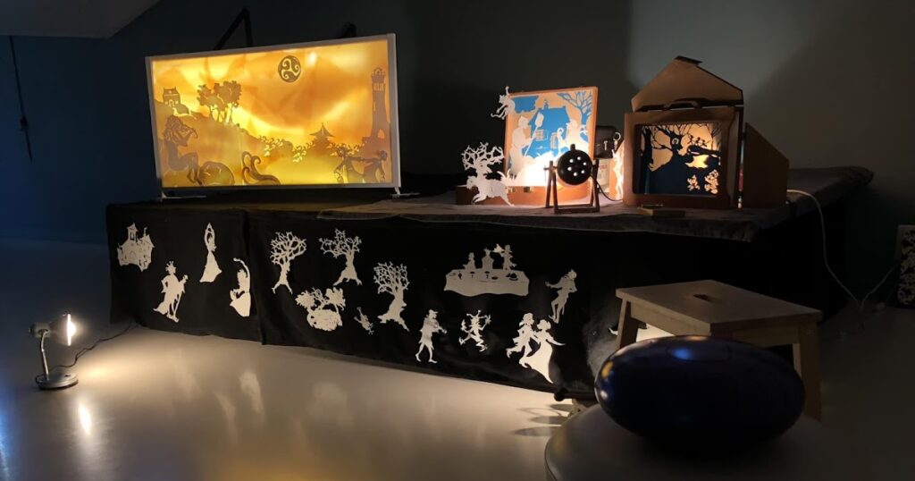 Scène de théâtre d’ombres avec des décors découpés éclairés, représentant des personnages et paysages féeriques.