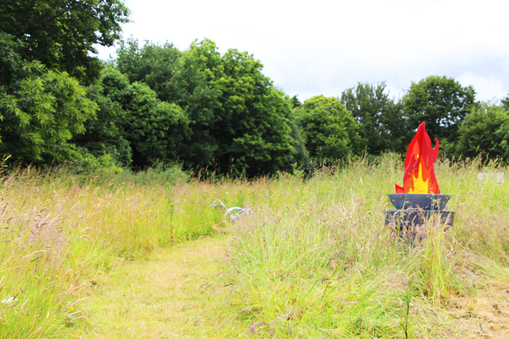 Une allée herbeuse traverse une prairie haute, avec une sculpture représentant une flamme rouge et jaune posée dans un support gris sur la droite.