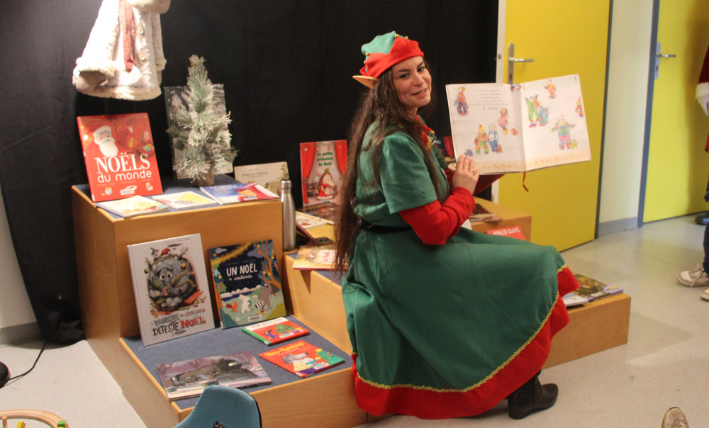 Une personne déguisée en lutin de Noël lit une histoire illustrée devant un présentoir de livres sur le thème de Noël.
