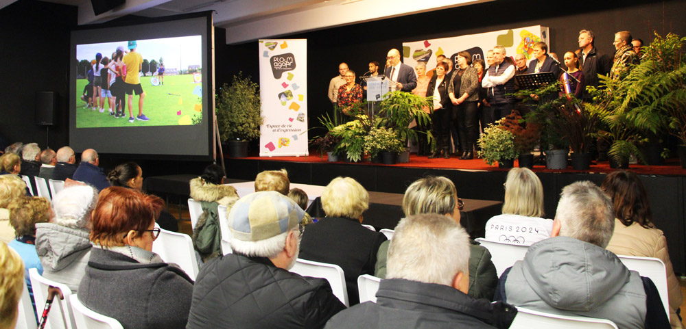Une assemblée est réunie dans une salle pour une cérémonie, avec un groupe d’élus et de représentants sur scène devant un pupitre, tandis qu’un écran projette une image d’activités sportives.