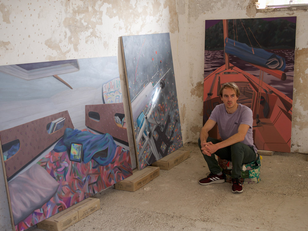 Un homme est accroupi dans une pièce aux murs abîmés, entouré de trois grandes toiles représentant des scènes d’intérieur de bateau et un voilier sur l’eau.
